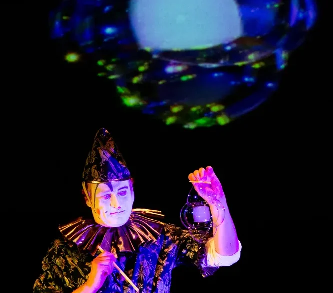 Un clown tient dans sa main une bulle de savon carrée, qui est également projetée sur un écran derrière lui.