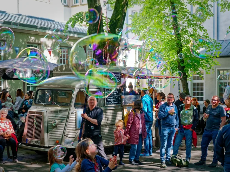 Une foule profite des bulles de savon devant une voiture ancienne, sous les arbres.