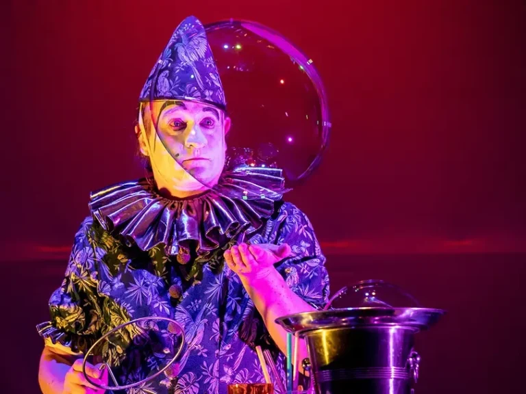 Artiste vêtue de vêtements colorés, coiffée d'un chapeau et tenant une bulle de savon géante sur fond rouge.
